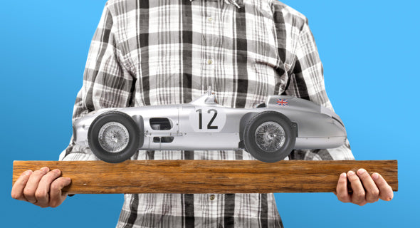 Person holding a Mercedes-Benz W196 R model kit on a wooden base with a blue background.